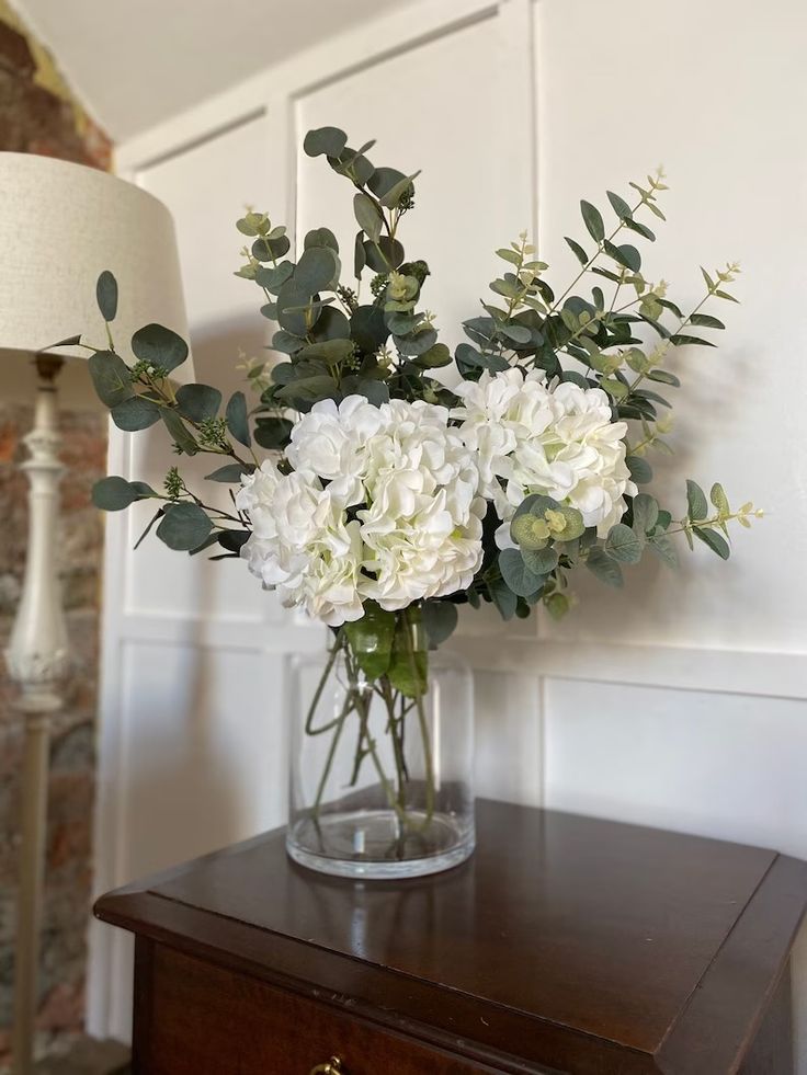 a vase filled with white flowers sitting on top of a wooden table next to a lamp