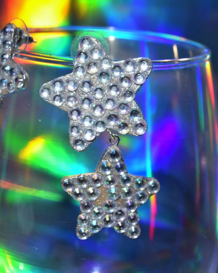 a pair of star shaped earrings sitting on top of a glass cup with colorful lights in the background