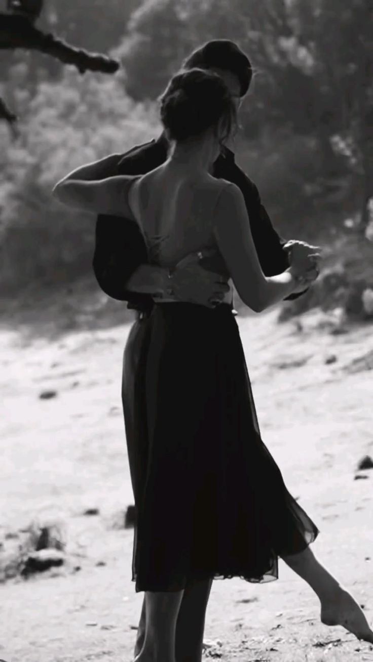 black and white photograph of two people dancing on the beach together, with trees in the background