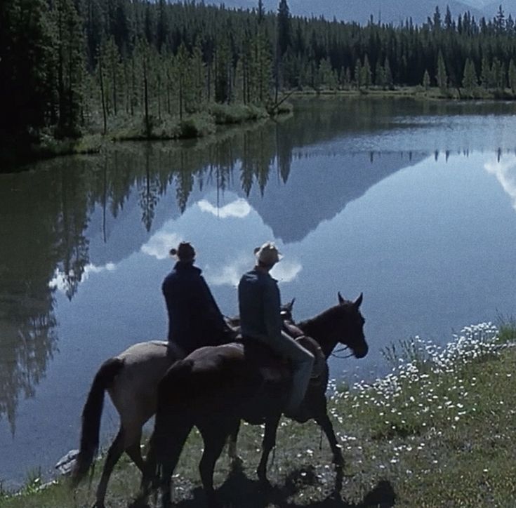 two people riding horses near a lake in the mountains with pine trees and clouds reflected in the water