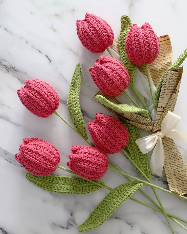 pink crocheted flowers with green stems on a marble surface, next to a ribbon