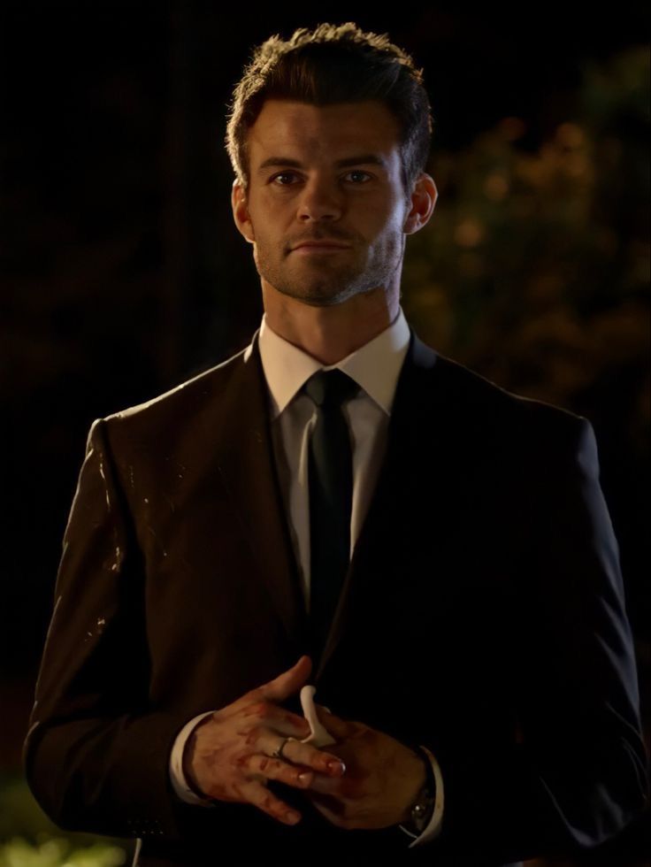 a man wearing a suit and tie standing in front of a dark background with his hands folded