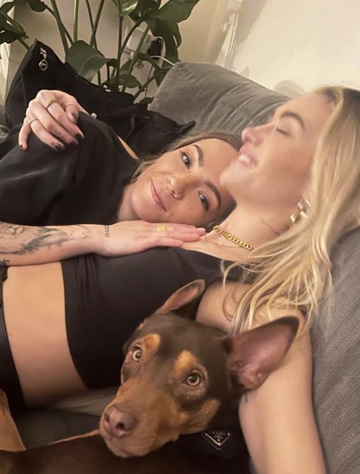 two beautiful women laying on top of a couch next to a dog