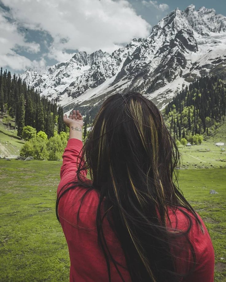 a woman in a red shirt flying a kite over a lush green field with snow covered mountains behind her