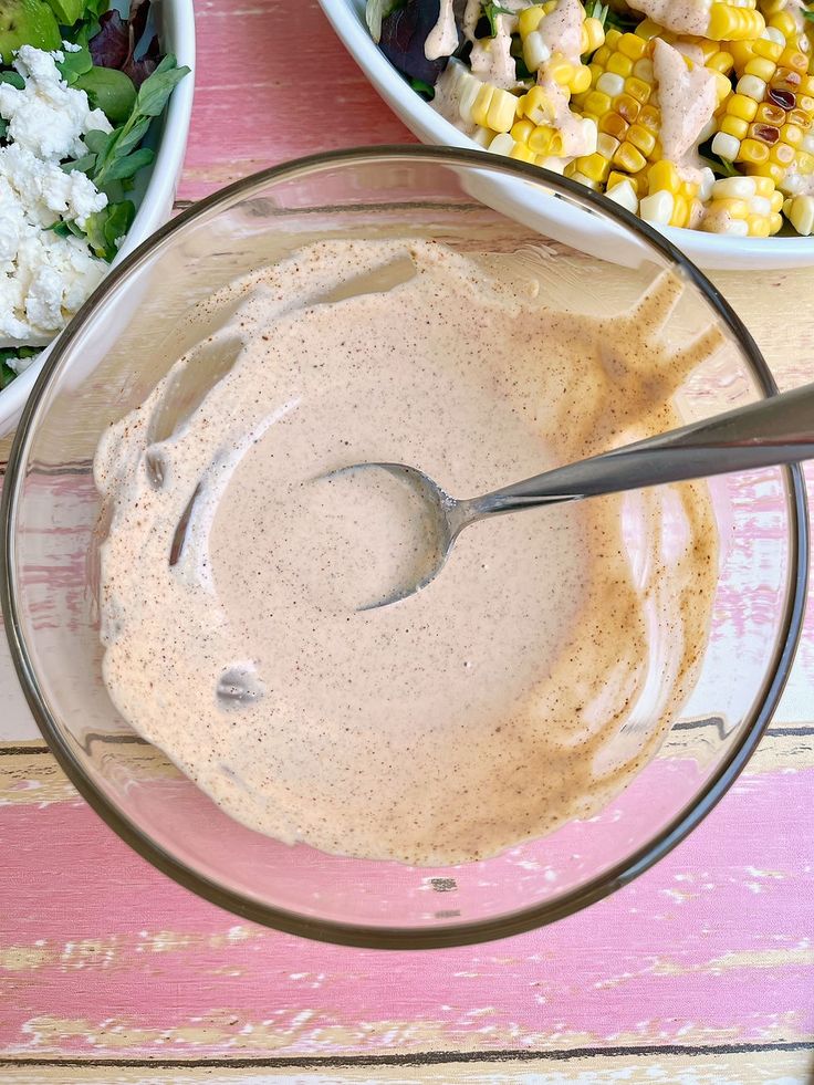 a bowl filled with dressing next to two bowls full of salad and corn on the side