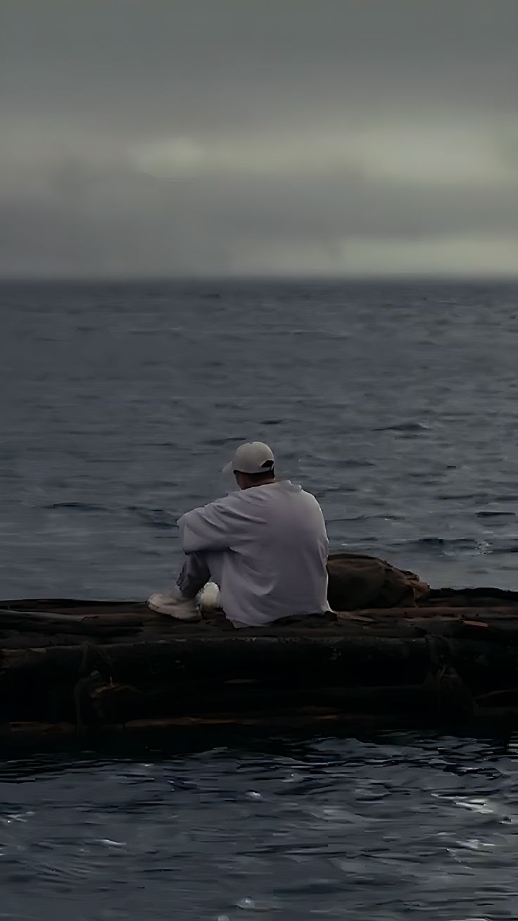 a man sitting on top of a wooden raft in the middle of the ocean under a cloudy sky