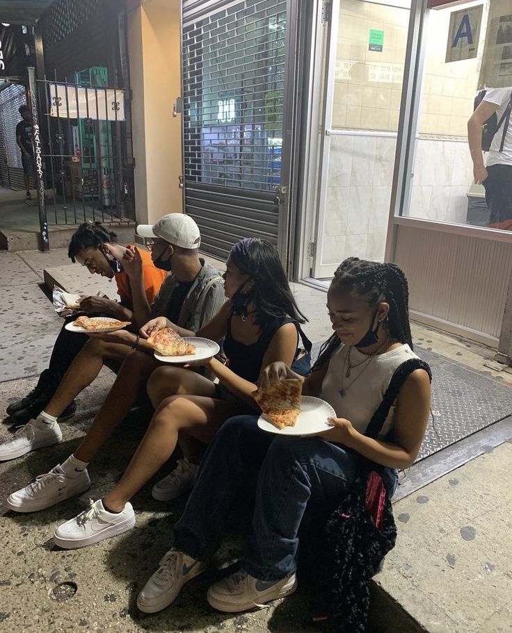 several people sitting on the curb eating pizza and talking to each other while holding plates