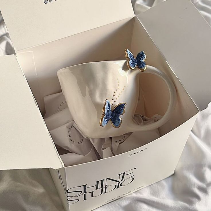 a white coffee cup with blue butterflies on it in a gift box, sitting on a bed