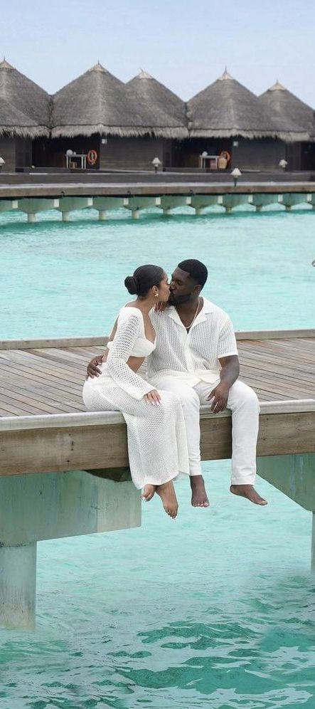 a man and woman sitting on a dock in front of some water with thatched huts