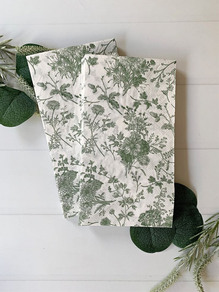 two napkins sitting on top of each other next to green leaves and plants in front of a white wall