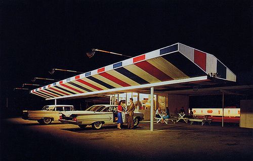 an old gas station at night with cars parked in the lot and people standing outside