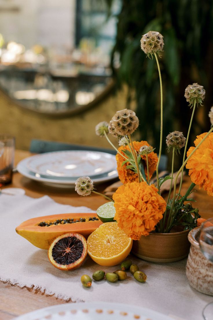 an arrangement of flowers and fruit on a table with plates, glasses and utensils