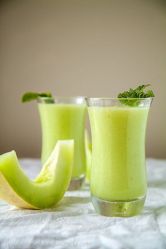 two glasses filled with green smoothie next to an apple slice on a white cloth