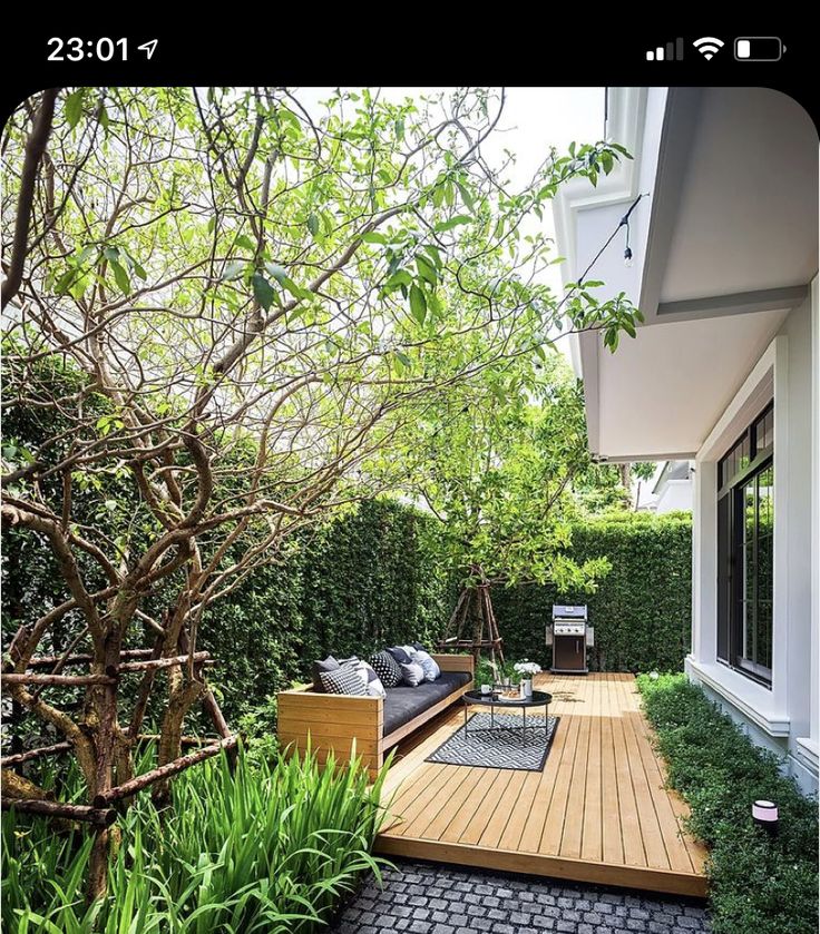 a wooden deck surrounded by greenery next to a white house with black furniture and windows