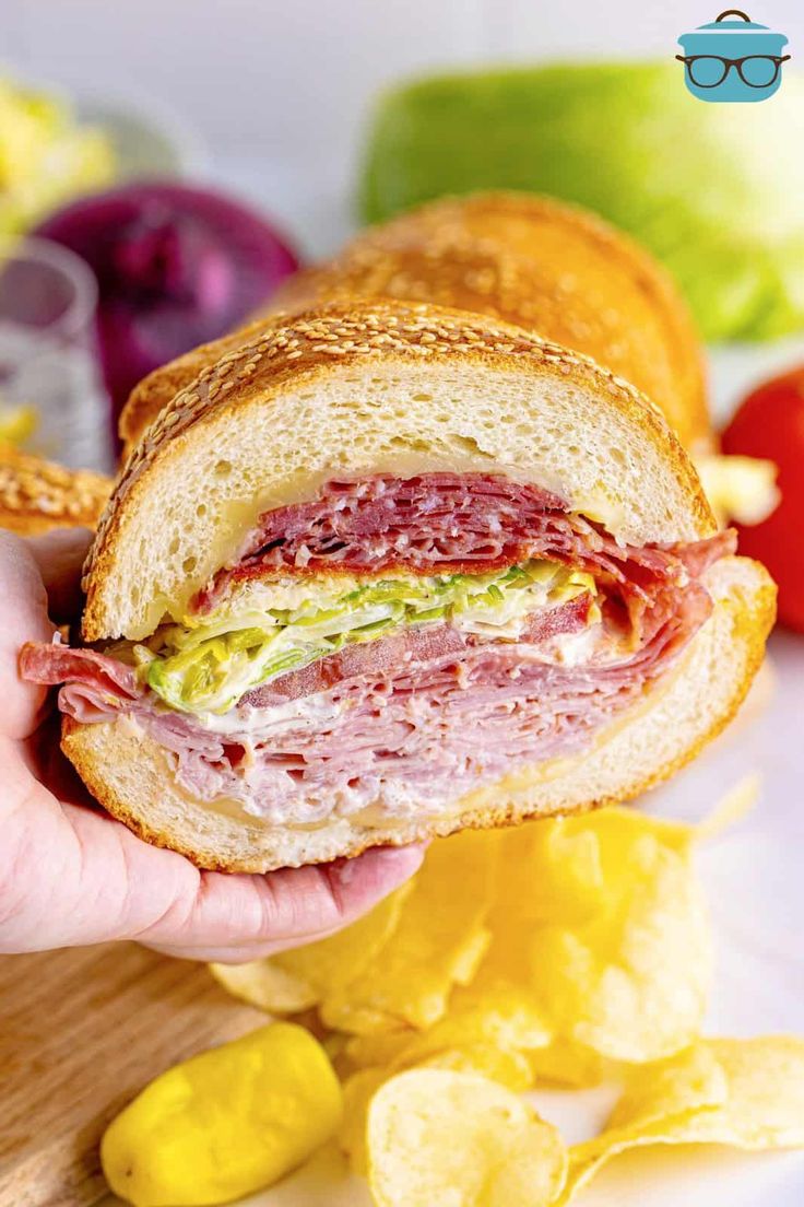 a person holding a sandwich in their hand with potato chips on the table next to it