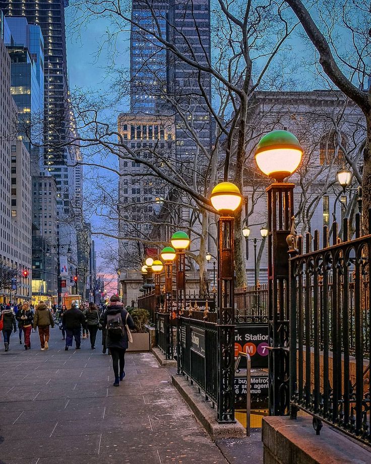 people are walking down the sidewalk in front of some tall buildings at night with lights on