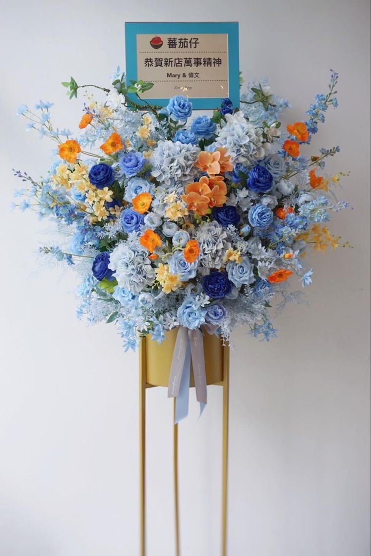 an arrangement of blue, orange and yellow flowers in a gold stand with a sign on it