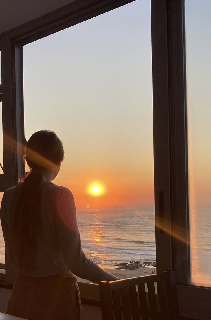 a woman standing in front of a window looking out at the ocean and sun setting