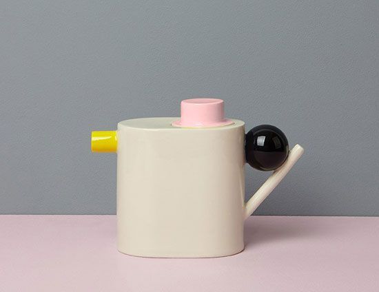 a white cup with a yellow handle and a black ball in the middle, sitting on a pink surface