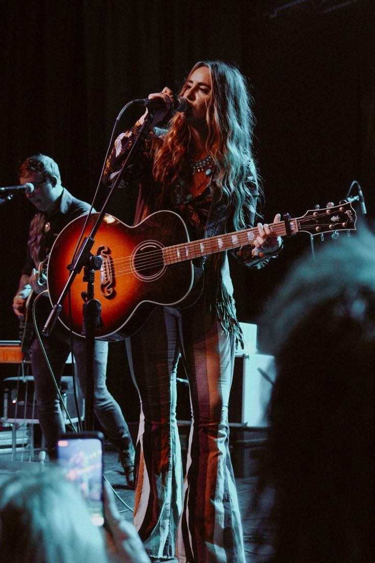 a woman singing into a microphone while holding an acoustic guitar in front of her on stage