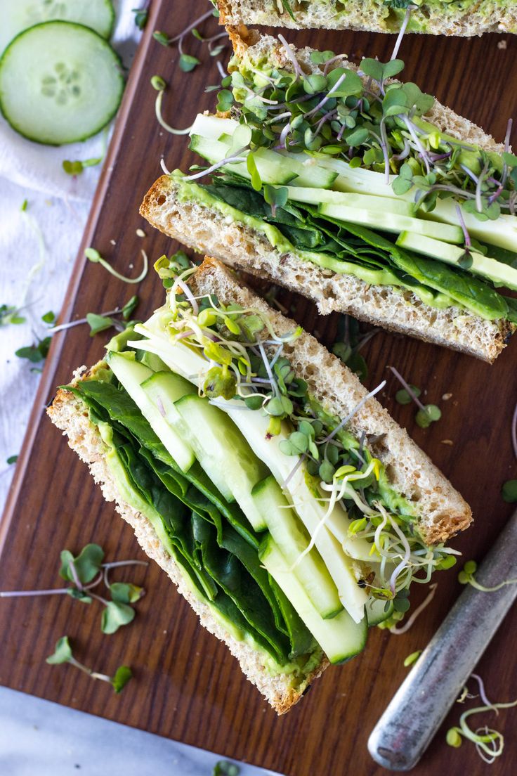 two slices of bread with cucumbers and sprouts on them, sitting on a cutting board