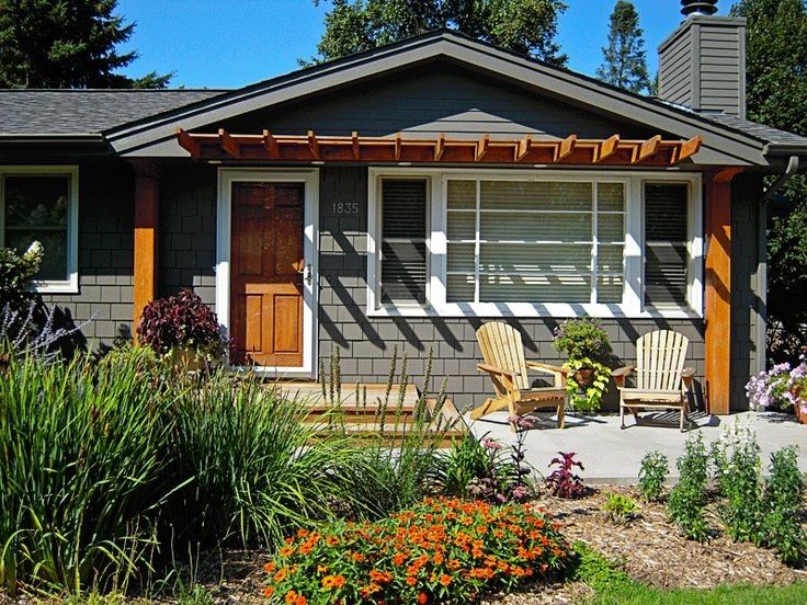 a small gray house with two chairs and flowers in the front yard, on a sunny day