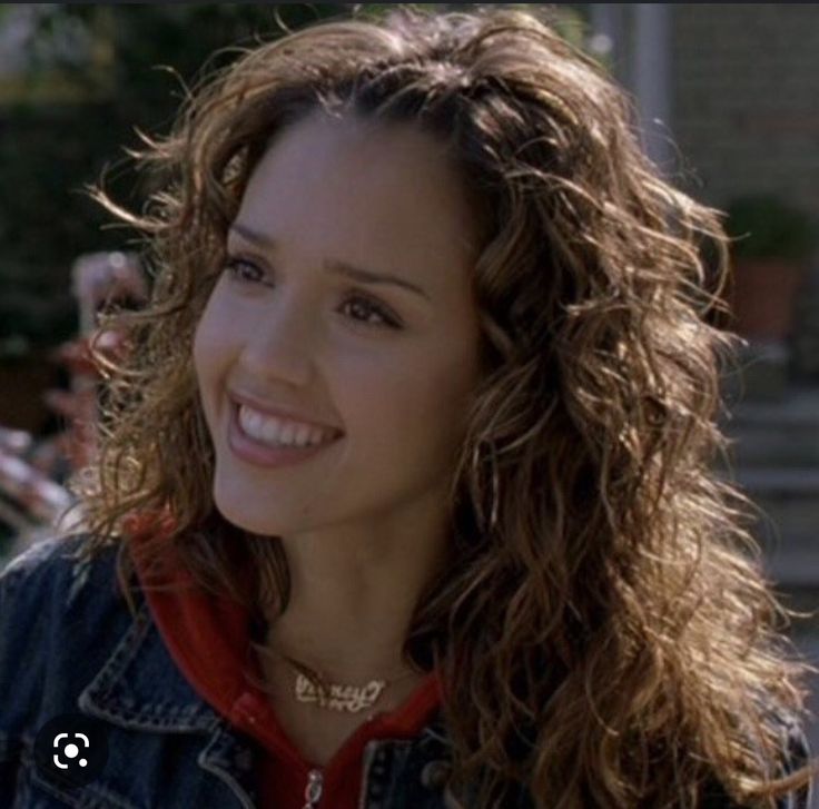 a woman with long curly hair wearing a jean jacket and smiling at someone in the background