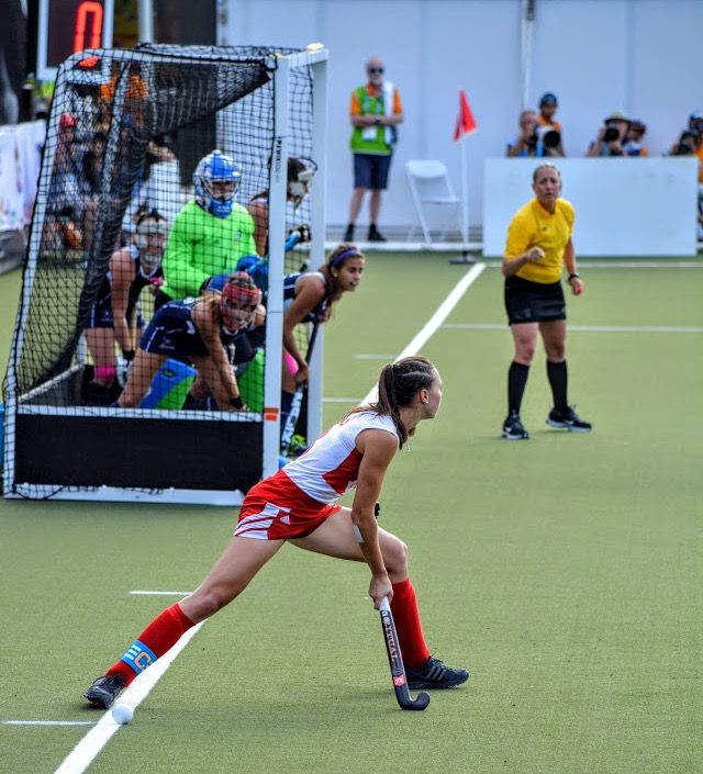 a woman playing field hockey on an outdoor court with people watching from the sidelines