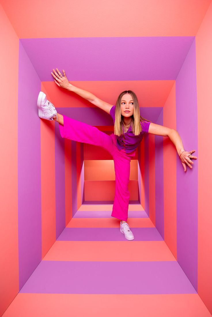 a woman in pink and purple poses with her arms spread out, while leaning against the wall