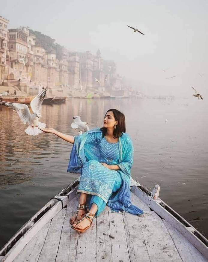 a woman sitting in a boat with seagulls flying over her and buildings on the shore