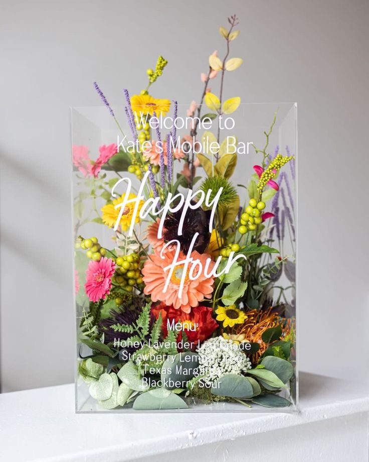 a clear acrylic sign that says happy hour with flowers and greenery in it