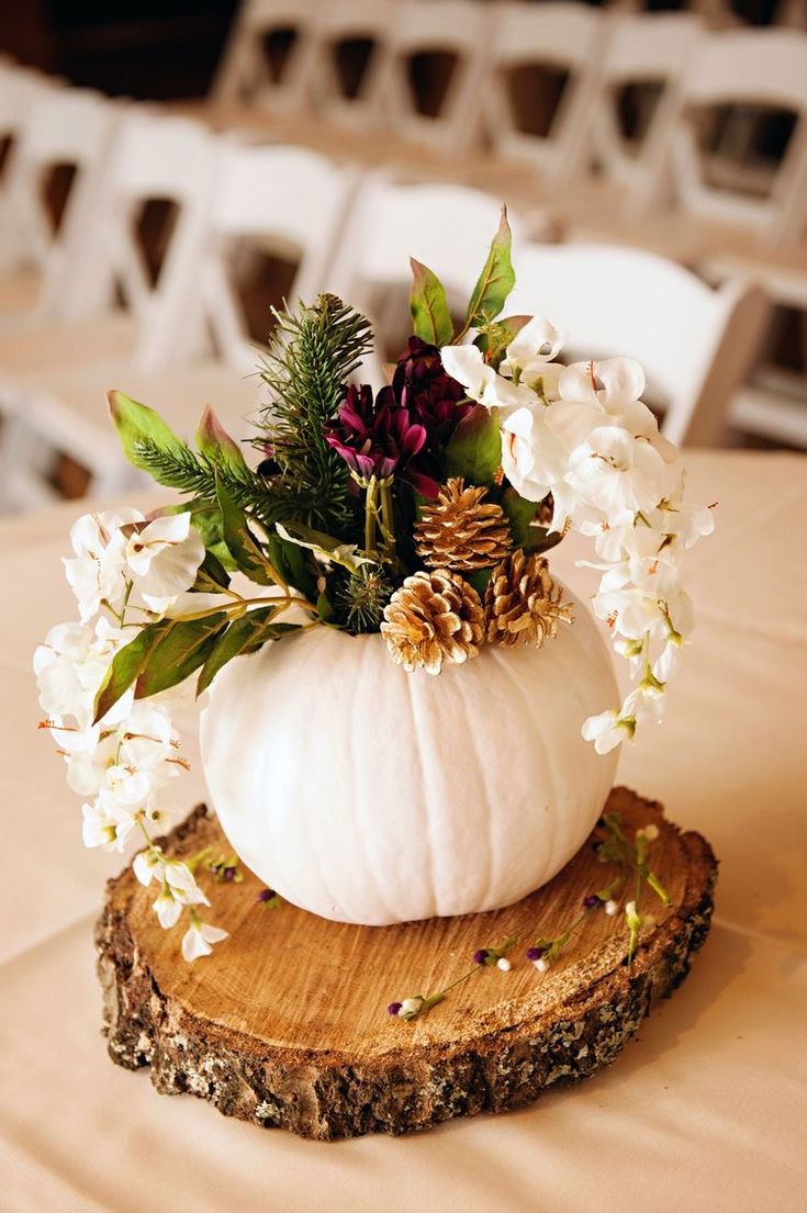 a white pumpkin decorated with greenery and pine cones sits on a wood slice at the head table