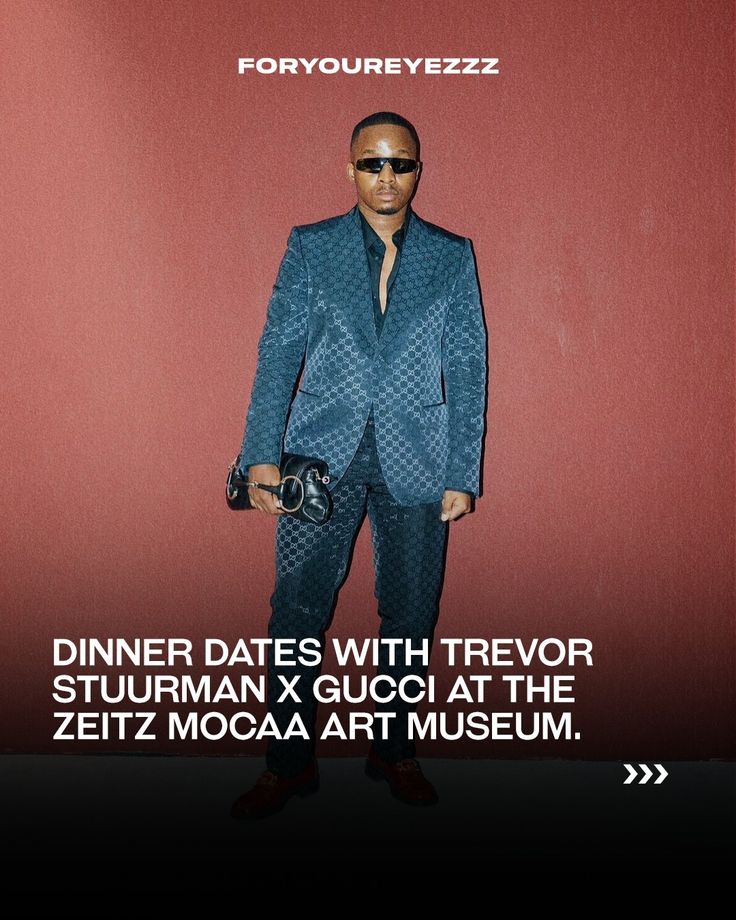 a man in a blue suit and sunglasses standing next to a red wall with the words dinner dates with trevor stuurman guco at the zetz moga art museum