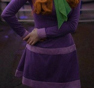 a woman with red hair wearing a purple dress and green bow tie standing in front of a building