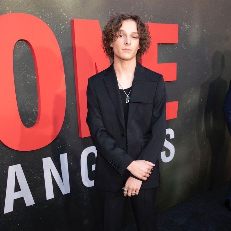 a young man in a black suit standing on a red carpet with his hands in his pockets