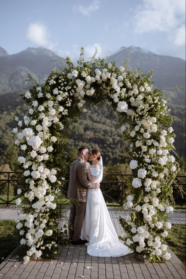 a bride and groom standing in front of a floral arch with white flowers on it