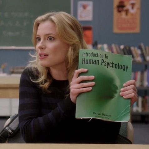 a woman sitting at a desk holding up a book in front of her face and looking off to the side