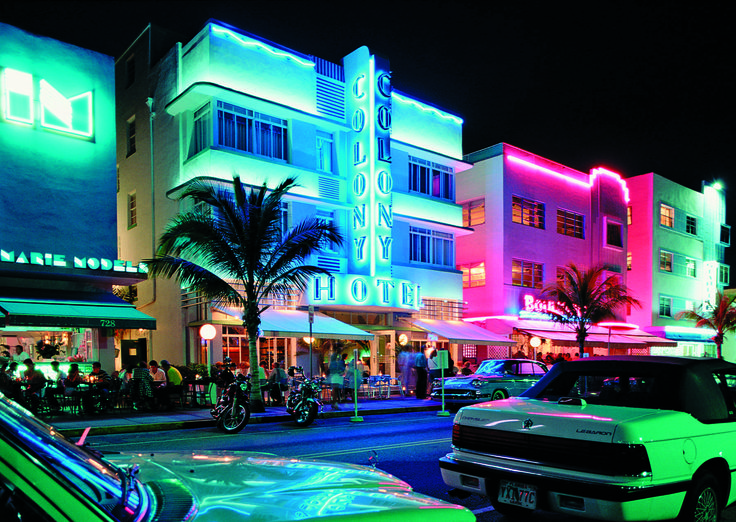 cars are parked on the street in front of a building with neon lights and palm trees