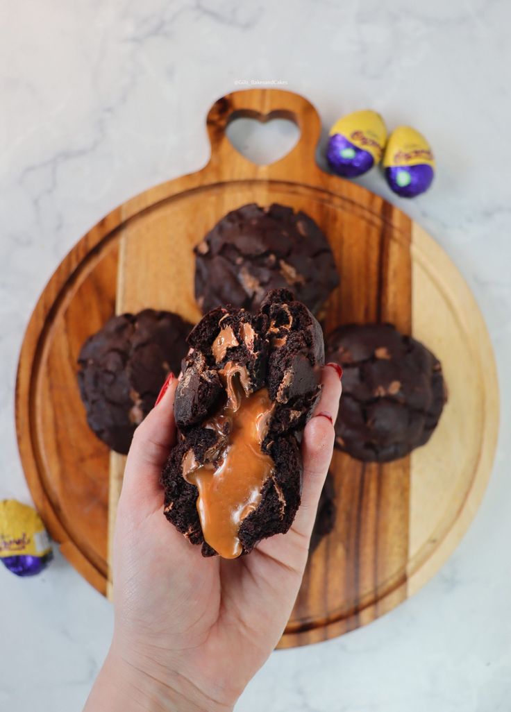 a person holding an egg stuffed cookie in front of some chocolate cookies on a wooden platter