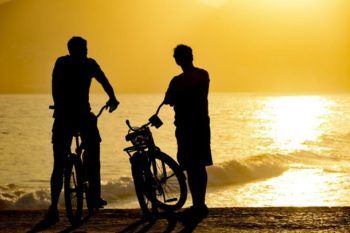 two people standing next to bikes on the beach at sunset with waves in the background