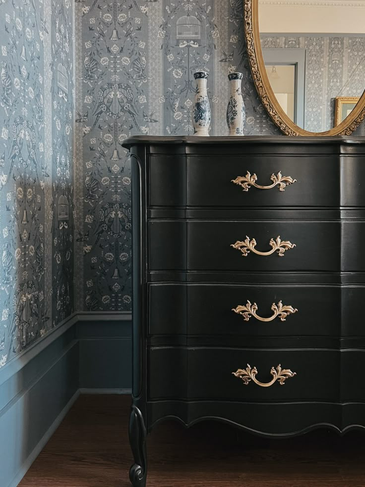 a black dresser with gold handles in front of a mirror and wallpapered walls