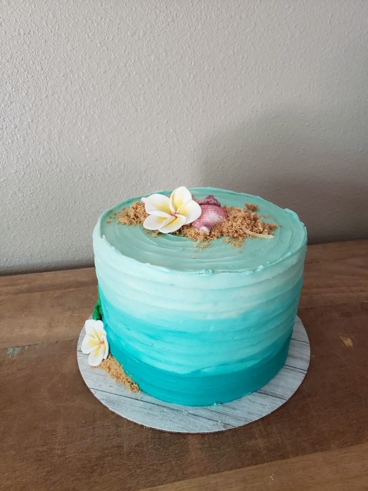 a blue cake with flowers and sand on top sitting on a wooden table next to a white wall