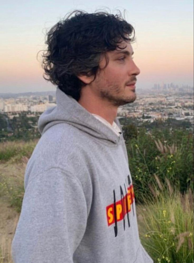 a man with curly hair standing in front of a cityscape and looking off into the distance