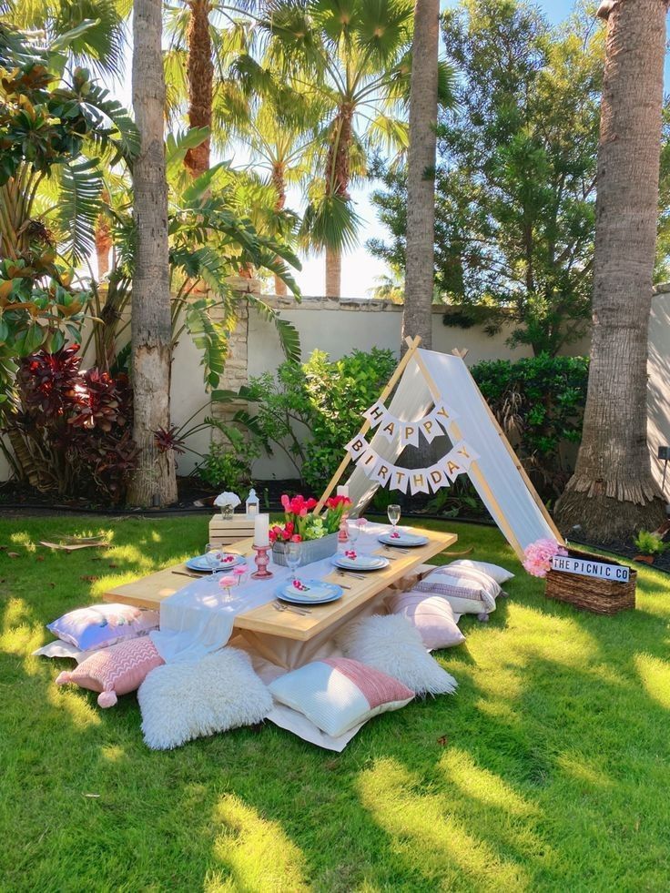 a picnic table set up in the grass with pink and white pillows on it, surrounded by palm trees