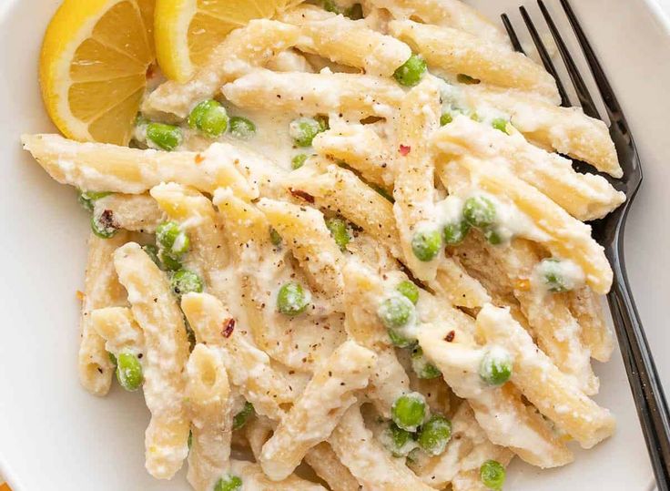 a white plate topped with pasta and peas next to a slice of lemon wedges