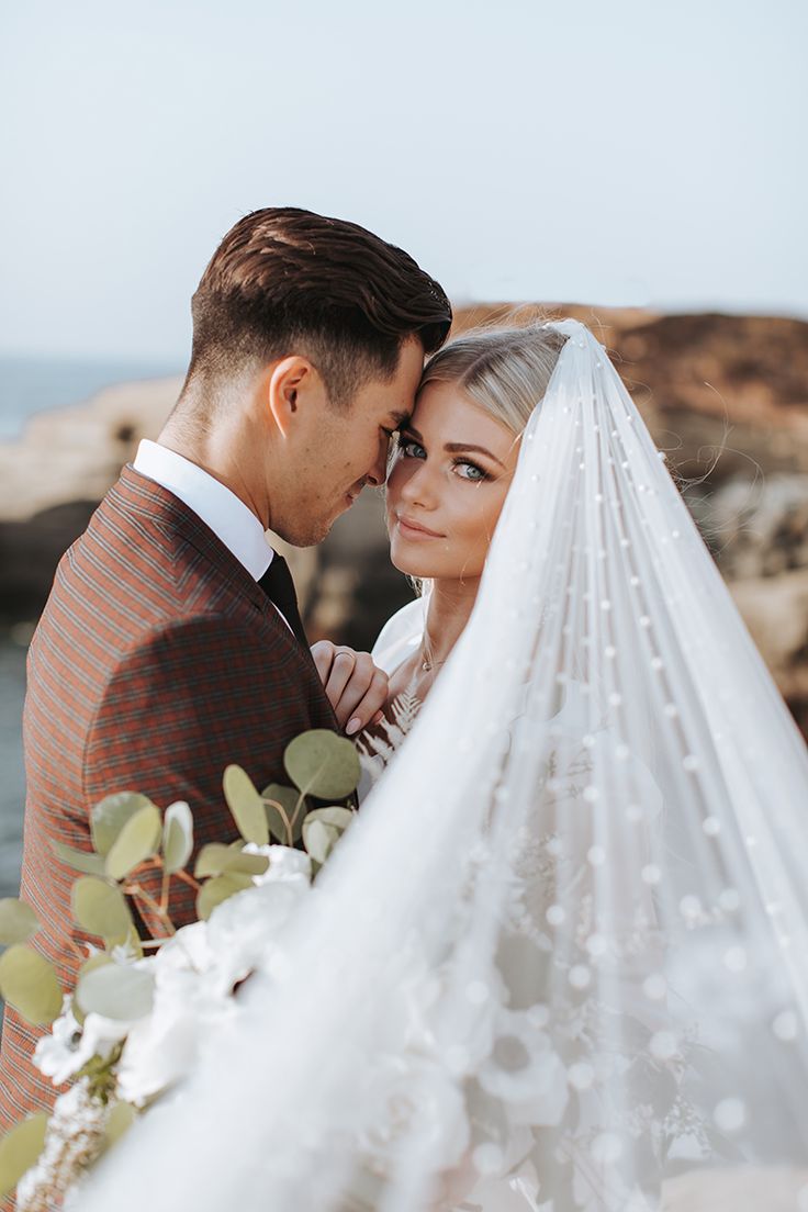 a bride and groom standing next to each other near the ocean with their veil blowing in the wind