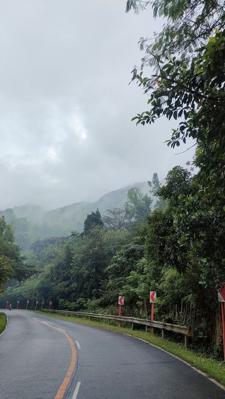 an empty road with trees and mountains in the background on a foggy day,