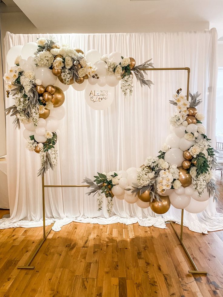 a white and gold balloon arch with flowers, greenery and balloons in the middle