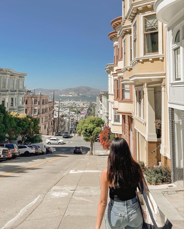 San Francisco painted ladies, antique buildings along the street facing a slanted hill. There’s a view of the city scape and a girl walking down the hill with the picture taken from her back. She has long black hair, jeans, and a trader joes tote bag. Sanfransisco Street Aesthetic, Cute Outfits For San Francisco, San Francisco Spring Break, Traveling To San Francisco, Sf Instagram Pictures, San Francisco Aesthetic Photos, San Francisco Astethic, Apartment In San Francisco, Life In San Francisco