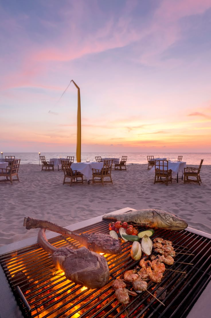 an outdoor grill on the beach at sunset with food cooking over it and chairs in the background